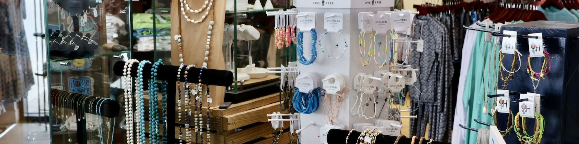 A jewelry and accessory store displays various necklaces, bracelets, and earrings on stands with clothes hanging in the background.