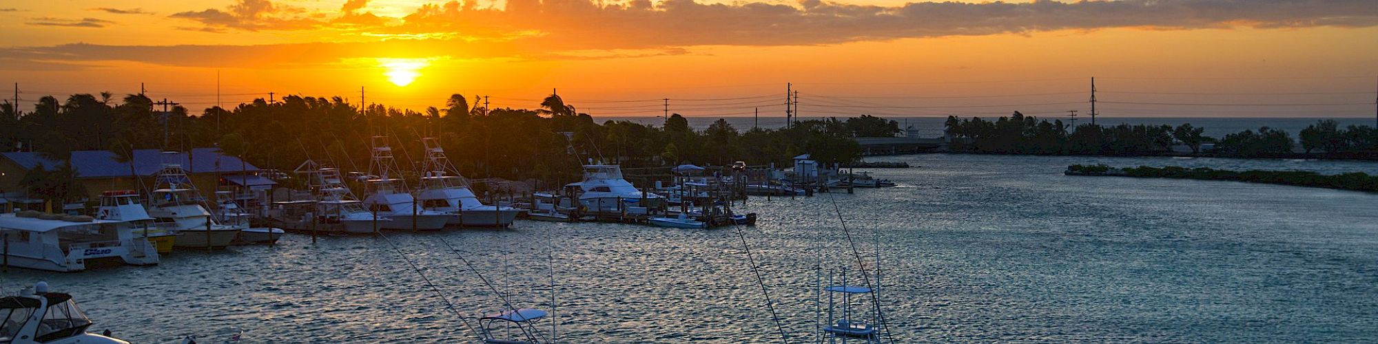 The image shows a serene marina at sunset with boats docked along a pier, calm waters, and a beautiful orange sky with the setting sun.