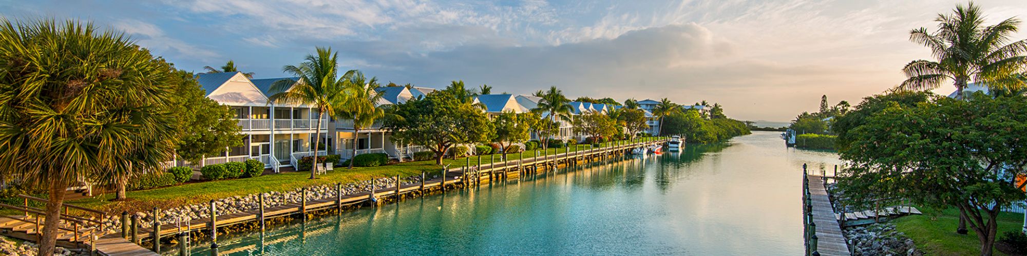 A serene waterfront scene features houses with docks, lush vegetation, and calm water under a blue sky with scattered clouds and soft sunlight.