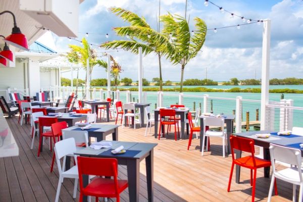 An outdoor restaurant with vibrant red and white chairs, wooden flooring, and a scenic waterfront view with palm trees and string lights.