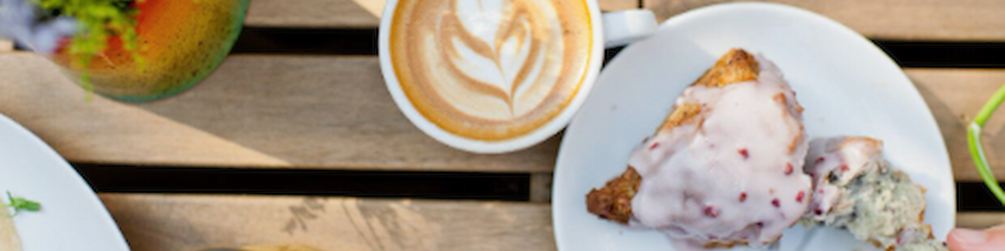 A latte with latte art, a scone with icing, a glass of juice, and a flower vase on a wooden table.