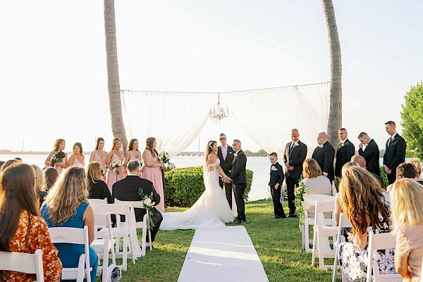 A wedding ceremony is taking place outdoors between two tall palm trees, with guests seated on either side of the aisle on white chairs, facing the couple.