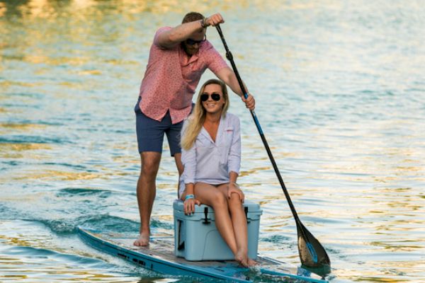 Two people are on a paddleboard; one is paddling while the other sits on a cooler. Both seem to be enjoying their time on the water, ending the sentence.