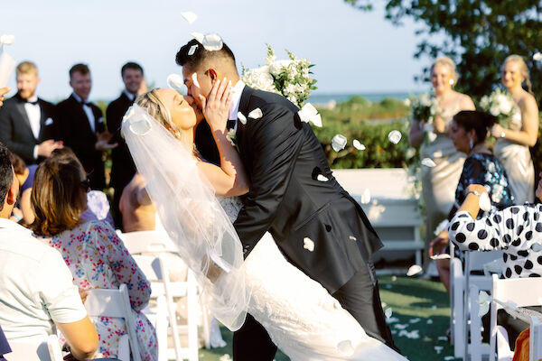 A couple is sharing a kiss at their wedding ceremony, surrounded by guests seated in white chairs, with an outdoor backdrop and floral decorations.