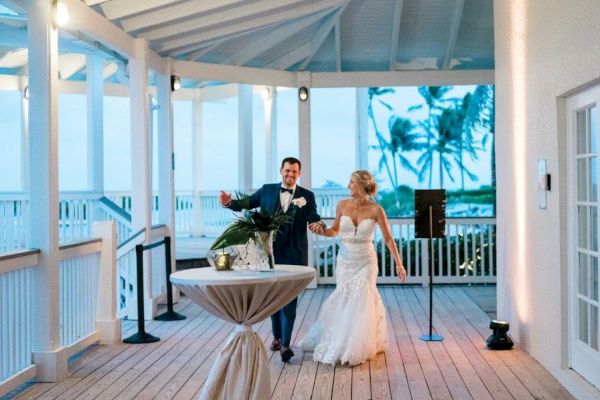 A bride and groom dance on a wooden deck under a pavilion with a beachy, tropical backdrop. The scene is joyful and festive, evident from their smiles.