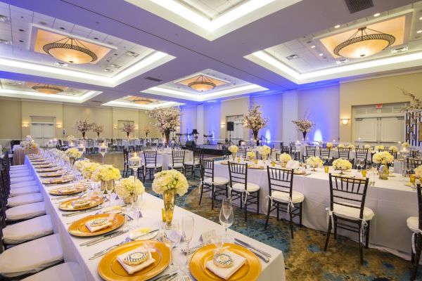 The image shows an elegantly set banquet hall with long tables, decorated with white and gold tableware, flower arrangements, and white chairs.