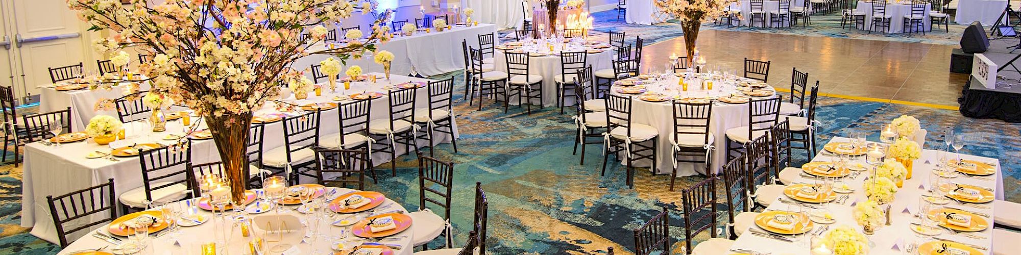 A beautifully decorated banquet hall is set for an event with round tables featuring floral centerpieces, and neatly arranged place settings.