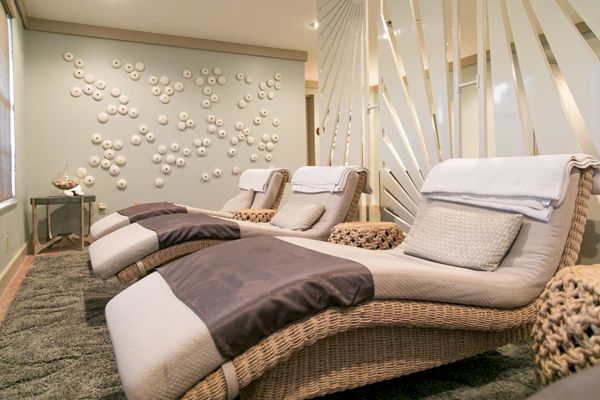 The image shows a serene spa room with three wicker lounge chairs, white towels, and plush gray cushions, set against a decorative wall display.