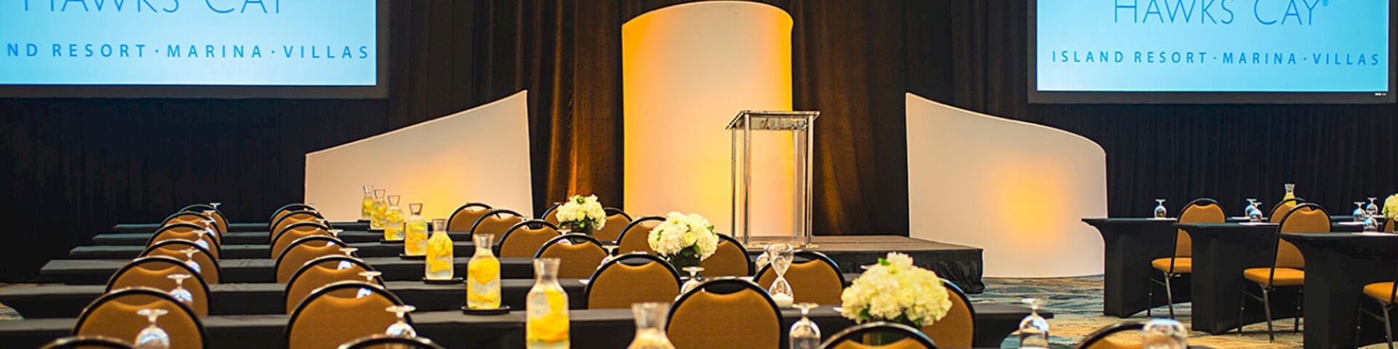 The image shows a conference room setup with rows of tables and chairs, a podium, and two projection screens displaying "Hawks Cay."