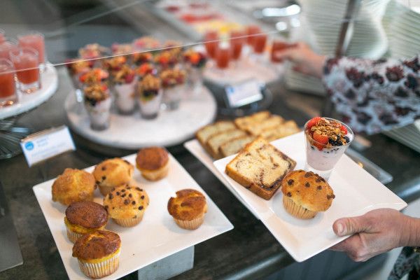 The image shows a buffet with muffins, pastries, and yogurt parfaits. A person's hand is holding a plate with muffins, bread, and a parfait.