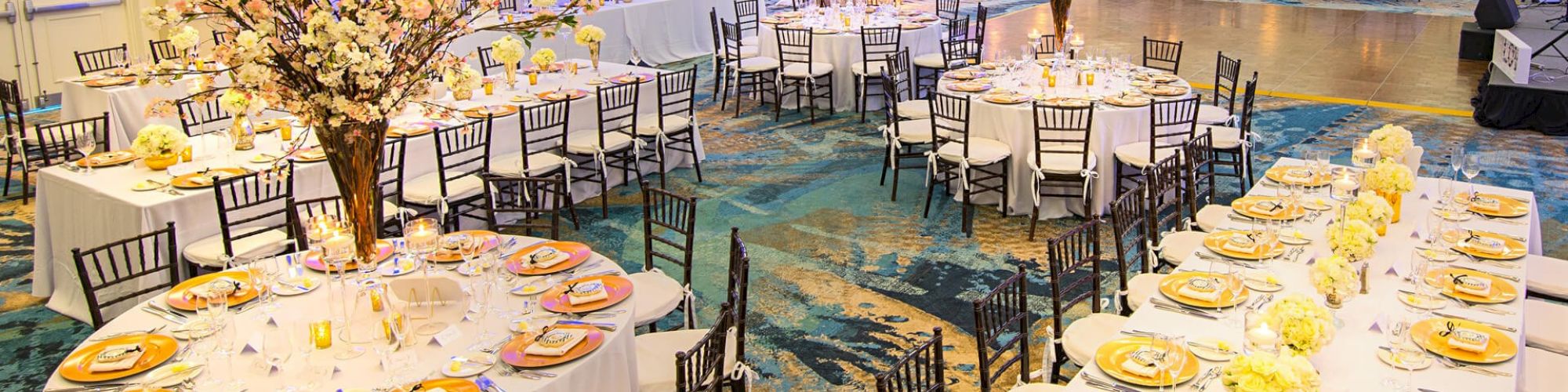 An elegant banquet hall is set for a formal event with round tables, floral centerpieces, and white and gold table settings, featuring cushioned chairs.