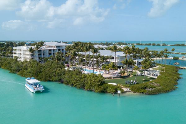 Aerial view of a tropical resort on a small, lush island with buildings, palm trees, pools, and a boat approaching the island on turquoise waters.