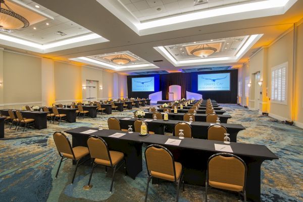 A conference room with rows of tables and chairs facing a stage, prepared with notepads, bottles, and pens, displaying two large screens.