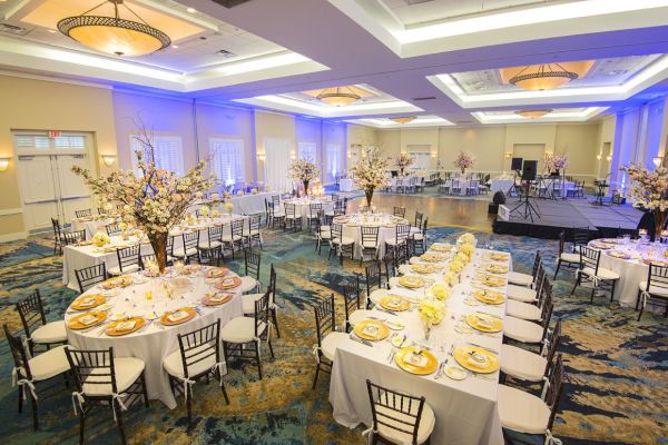 A decorated banquet hall with round tables set with plates and flowers, along with a stage and blue lighting is seen in the image.