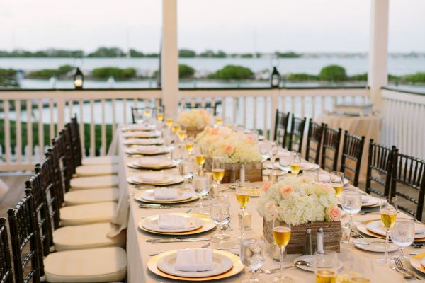 An elegantly set long dining table with white plates, gold chargers, and floral centerpieces on a covered patio with a view of the water in the background.