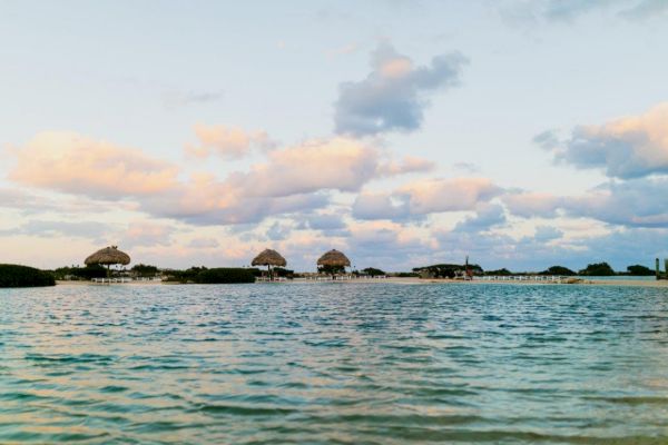 The image shows a serene beach scene with umbrellas and buildings in the background, under a partly cloudy sky with water in the foreground.