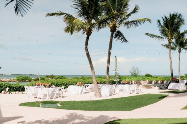 Outdoor event setup with white tables and chairs, tropical palm trees, string lights, and an ocean view in the background, with clear skies.