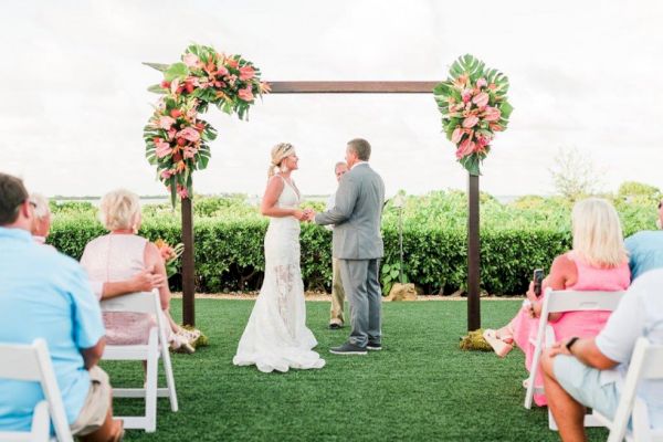 A couple is getting married outdoors under a floral arch, exchanging vows. Guests are seated, watching the ceremony on a grassy area.