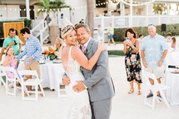 A bride in a white gown and a groom in a gray suit dance outdoors at a wedding reception with guests seated at tables around them.