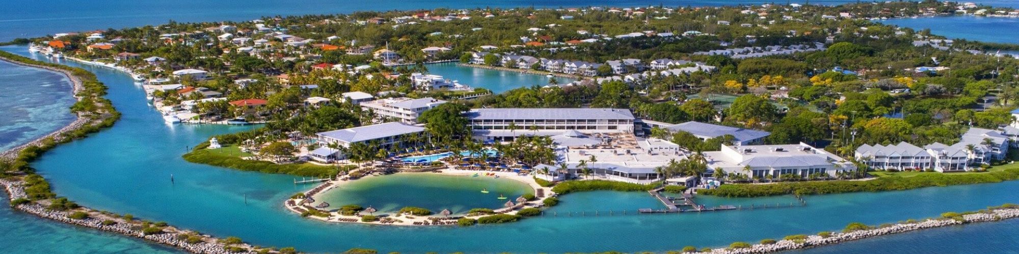 An aerial view of a resort on a small island surrounded by turquoise waters and connected land masses, showing buildings, pools, and greenery.