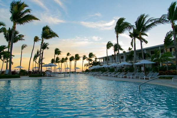 A serene poolside scene at a resort features lounge chairs, parasols, palm trees, and a modern building at sunset, creating a relaxing ambiance.