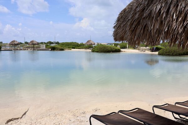 A tranquil beach scene with clear water, a thatched umbrella, lounge chairs, and lush greenery under a partly cloudy sky.