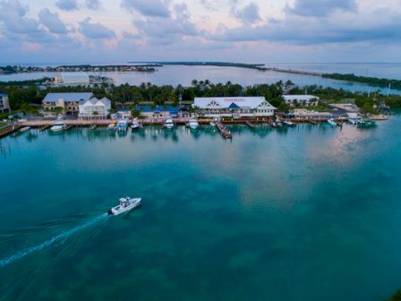 An aerial view of a coastal town with buildings along the shoreline, clear turquoise water, a boat cruising, and a bridge in the background.