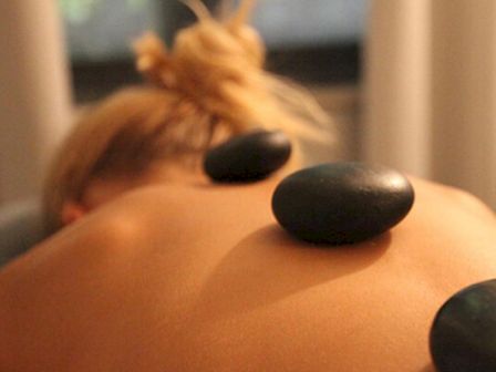 The image shows a person receiving a hot stone massage, with three smooth black stones placed along their back, in a relaxing setting.