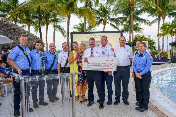 A group of people, including rescue personnel in uniform, standing together outdoors, holding a ceremonial check in front of a podium and poolside.