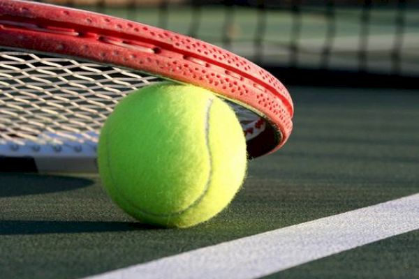 A tennis ball on a court with a racket lying over it, near the net and the court's white boundary line.