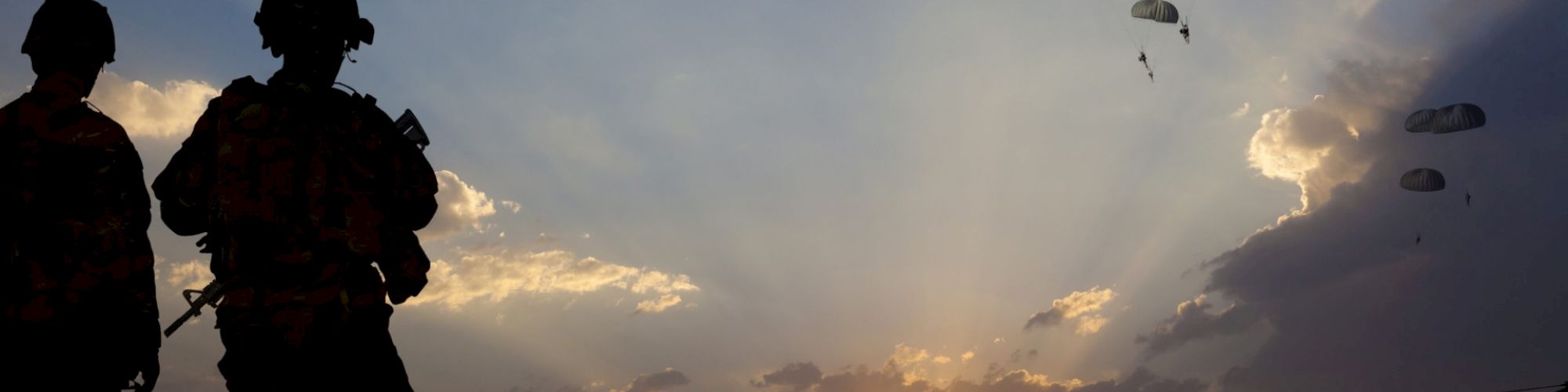 Silhouettes of soldiers with a dramatic sunset backdrop, parachutes dropping from the sky, and a helicopter in flight.