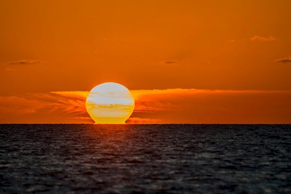 A stunning sunset over a calm ocean with a large, glowing orange sun partly obscured by clouds and reflecting off the water surface.