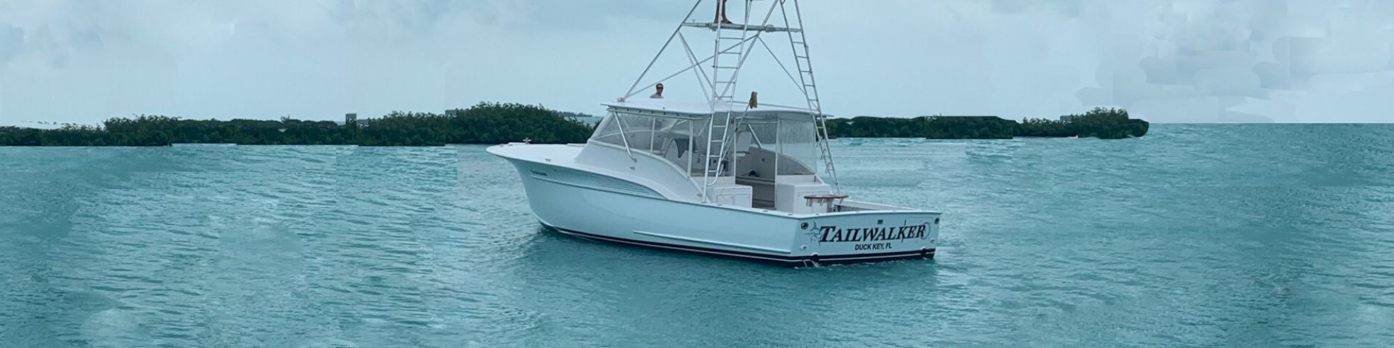 A white boat named "Tailwalker" is anchored in calm, turquoise waters with a distant shoreline and cloudy sky in the background.