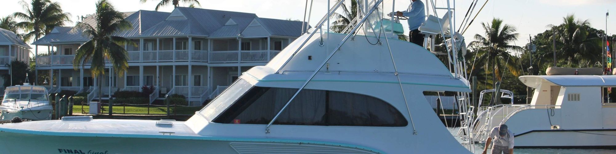 A white yacht named "Final Final" is docked with two people on board, set against a background of palm trees and multi-story buildings.