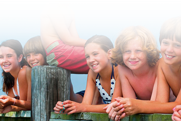 The image shows a group of five children leaning on a wooden fence, smiling and enjoying a sunny day at what appears to be a beach or poolside.