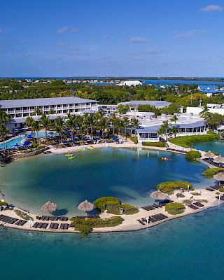 A coastal resort with multiple buildings, a private lagoon, loungers on the beach, and lush greenery. Clear blue water surrounds the area.