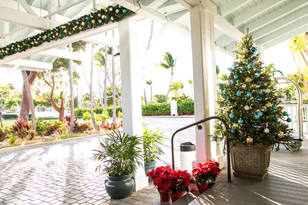 A festive outdoor setting with a decorated Christmas tree, garlands, potted plants, and poinsettias, under a covered walkway on a sunny day.