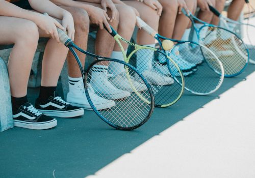 Several people seated on a bench, holding tennis rackets, with their legs and rackets visible. They all wear different types of shoes.