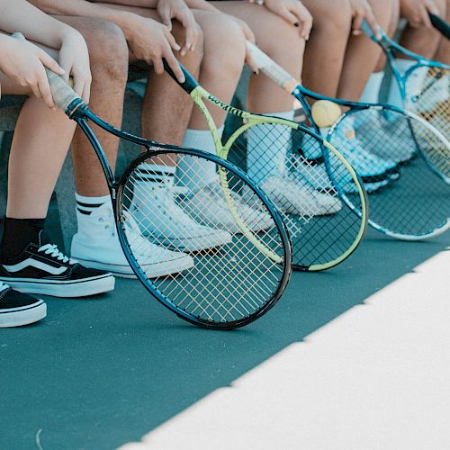 Several people seated on a bench, holding tennis rackets, with their legs and rackets visible. They all wear different types of shoes.