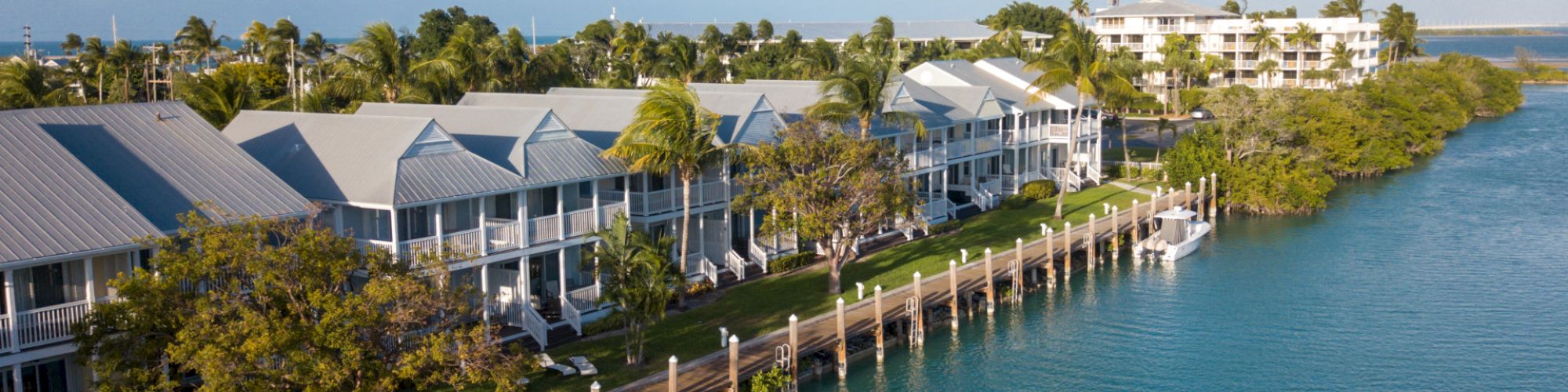 A waterfront scene with several coastal houses, lush greenery, and a dock extending into a calm blue waterway, under a partly cloudy sky.