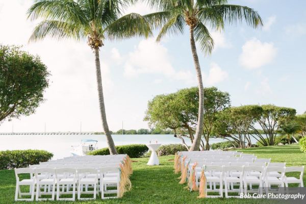 Outdoor wedding setup with rows of white chairs under palm trees, overlooking a scenic water view and a bridge in the background.