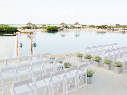 The image shows an outdoor wedding setup with white chairs arranged in rows facing a decorated arch, overlooking a serene body of water.