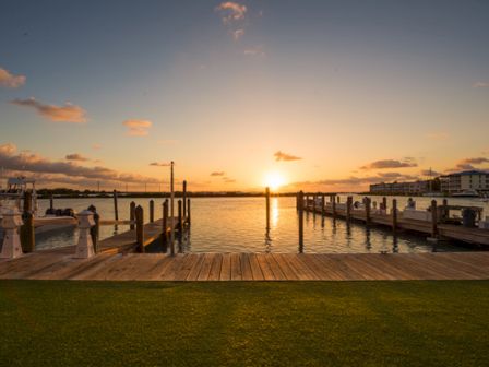 A serene sunset view over a harbor, with docks and boats, complemented by a wooden boardwalk and lush green foreground.