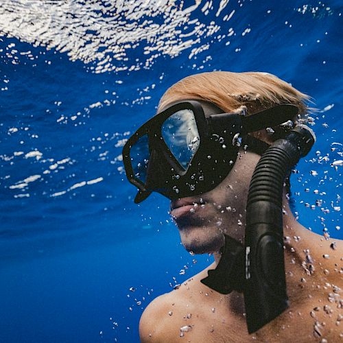 A person is underwater, wearing a snorkel mask and looking upward. Bubbles and reflections are visible in the surrounding blue water.