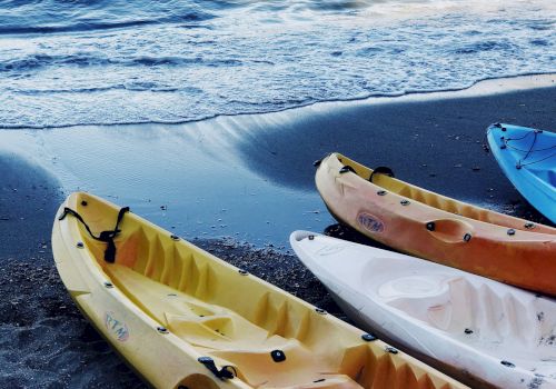 Several kayaks are lined up on a sandy beach near the shoreline, with gentle waves from the ocean in the background.