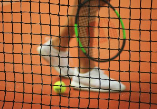 The image shows a tennis racket, a tennis ball, and a player's legs on a clay court viewed through a tennis net.