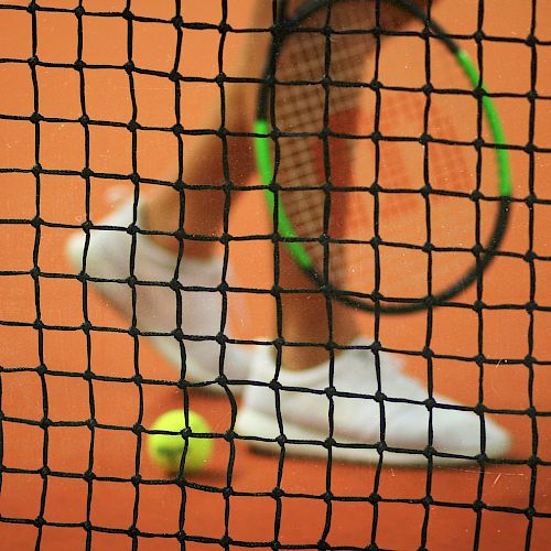 The image shows a tennis racket, a tennis ball, and a player's legs on a clay court viewed through a tennis net.