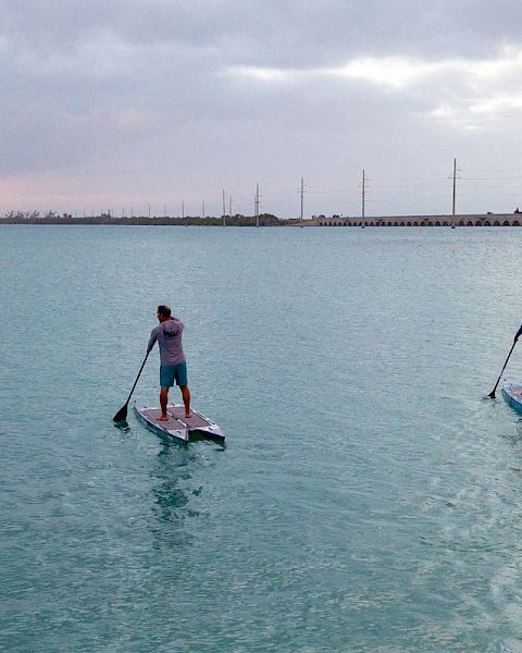 Two people are stand-up paddleboarding on a large body of water at sunset with a long bridge and some trees in the distance.