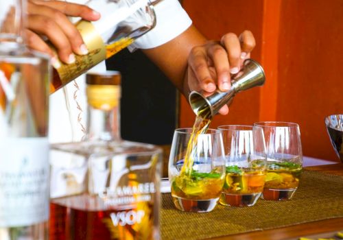 A person is pouring liquid from a jigger into glasses next to various bottles, likely preparing drinks on a bar counter.