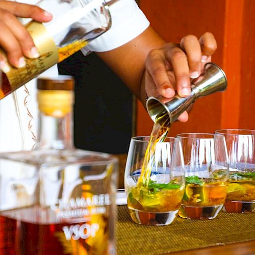 A person is pouring liquid from a jigger into glasses next to various bottles, likely preparing drinks on a bar counter.